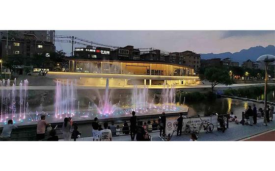 Visitors are enjoying an illuminated fountain on the riverside in the evening.