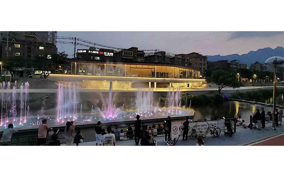 Visitors are enjoying an illuminated fountain on the riverside in the evening.