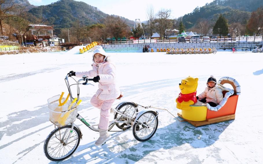 a girl riding a tricycle is pulling a sled with another girl on.