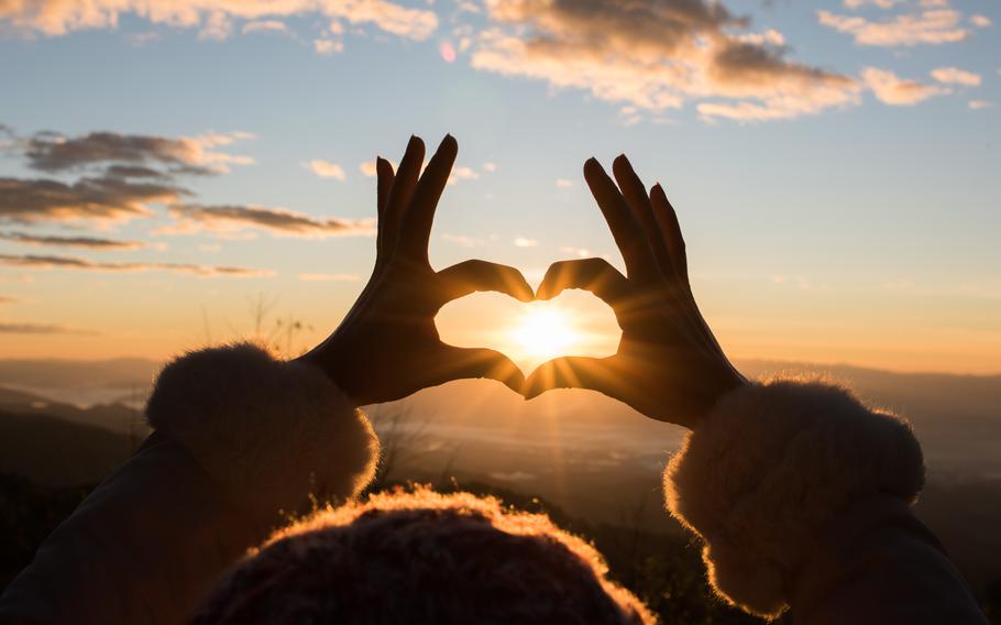 a person making a heart shape by using their fingers and the sunrise can be seen inside the shape.